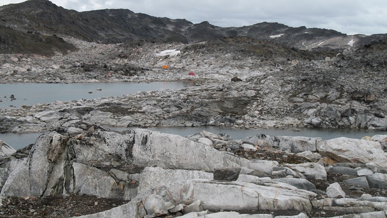 Et landskab af klipper og vand ses. Det er Manitsoq-strukturen i Grønland. Der er et rødt og et orange telt i baggrunden. Det er feltlejren.